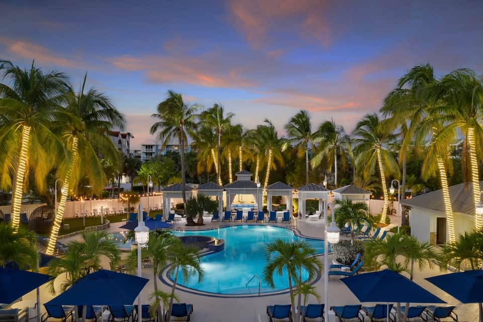 Resort pool at dusk with illuminated palms, curved pool design, blue lounge chairs, and white poolside pavilion structures