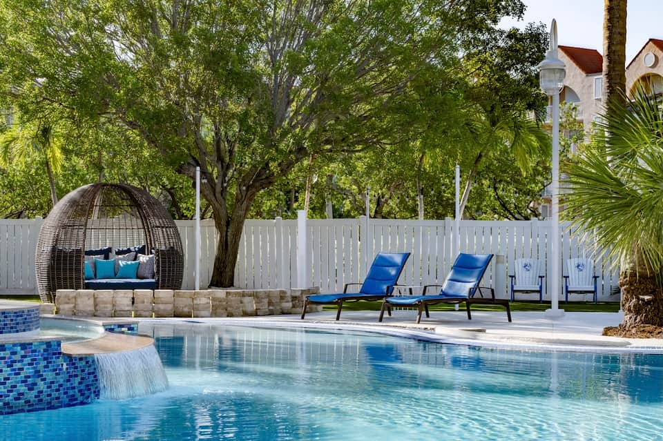 Luxury pool with waterfall feature, woven hanging chair, blue lounge chairs, white fence, and mature shade trees