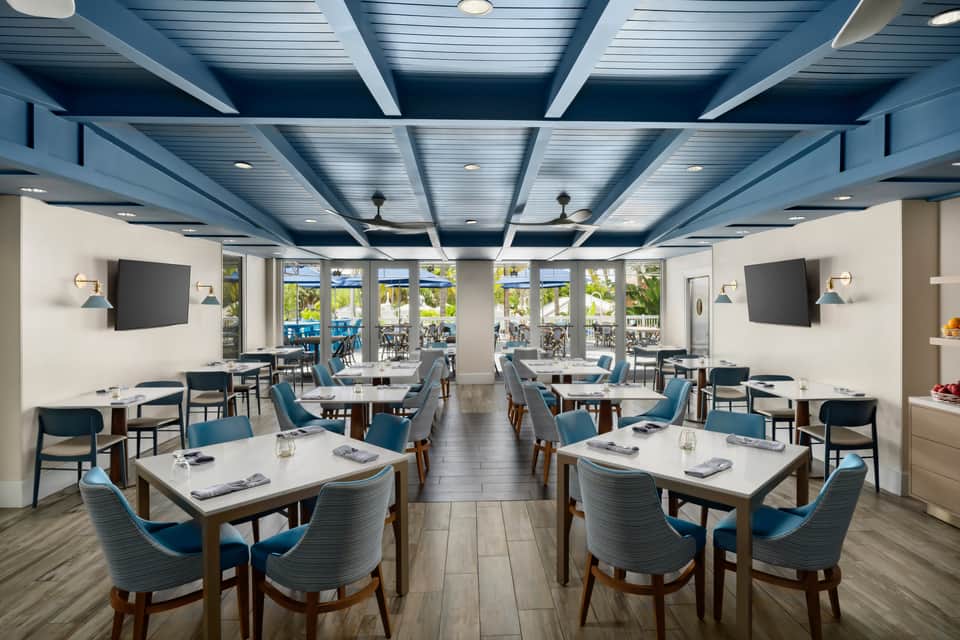 Modern restaurant dining room with blue chairs, white tables, lattice ceiling, and ocean views through glass doors