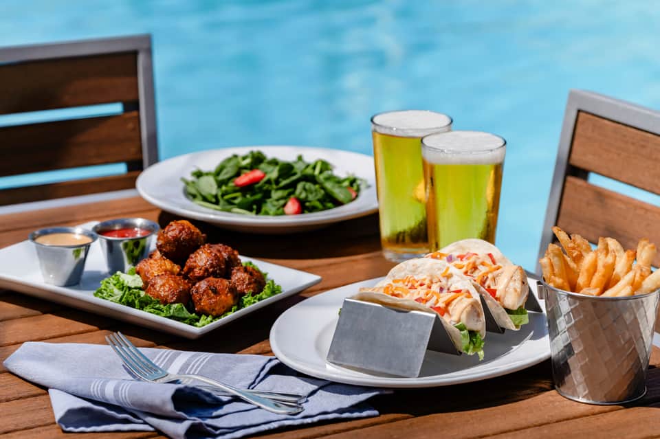 Poolside dining table with food, drinks, and ocean view; tacos, meatballs, salad, and beer