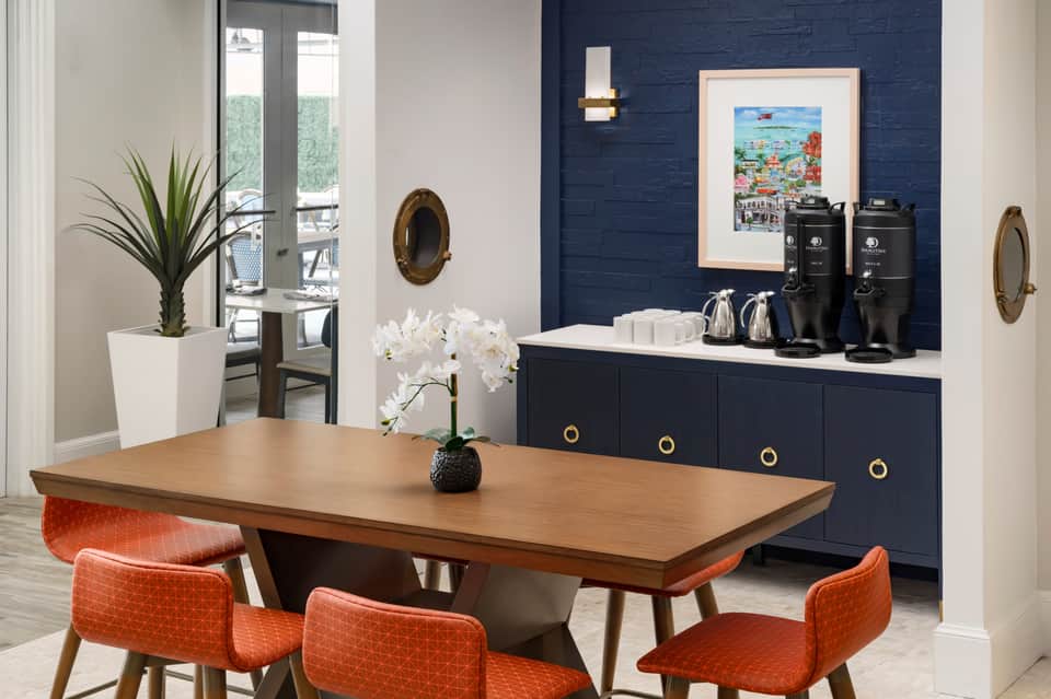 Modern hotel foyer with navy accent wall, beverage station, and wood dining table with orange chairs