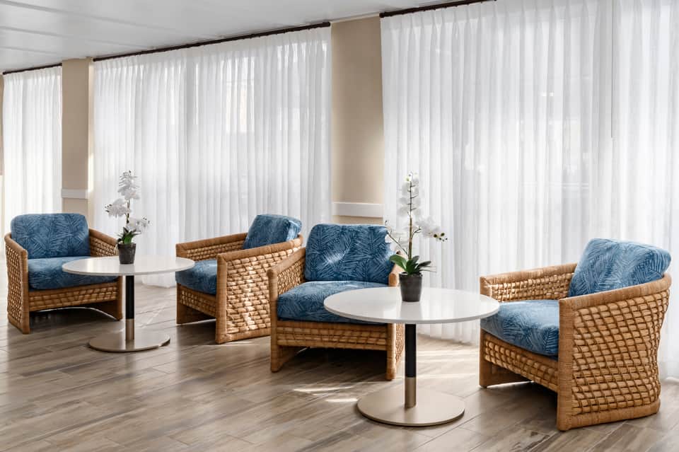 Bright lounge area with wicker chairs, blue patterned cushions, white sheer curtains, and round tables