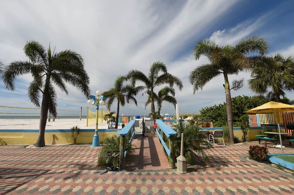 Beachfront resort grounds with palm trees, blue pathway bridge, playground, and yellow umbrellas