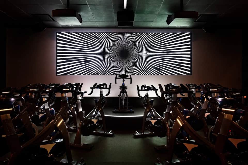 Fitness studio with rows of stationary bikes and large geometric light display wall