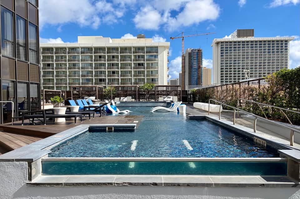 Rooftop pool with blue lounge chairs, city skyline, and modern high-rise buildings