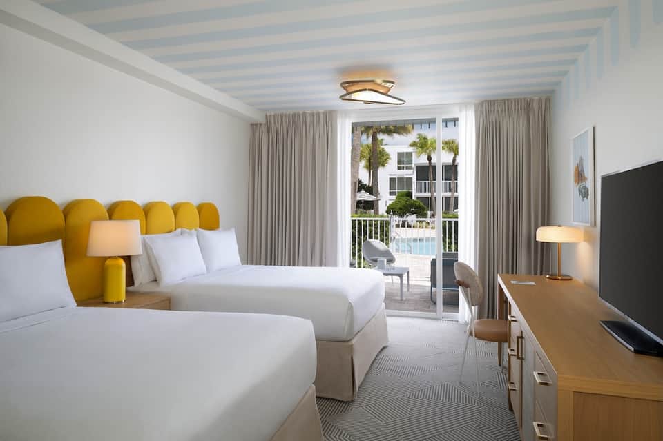 Bright hotel bedroom with twin beds, yellow headboard, ocean view balcony, and striped ceiling
