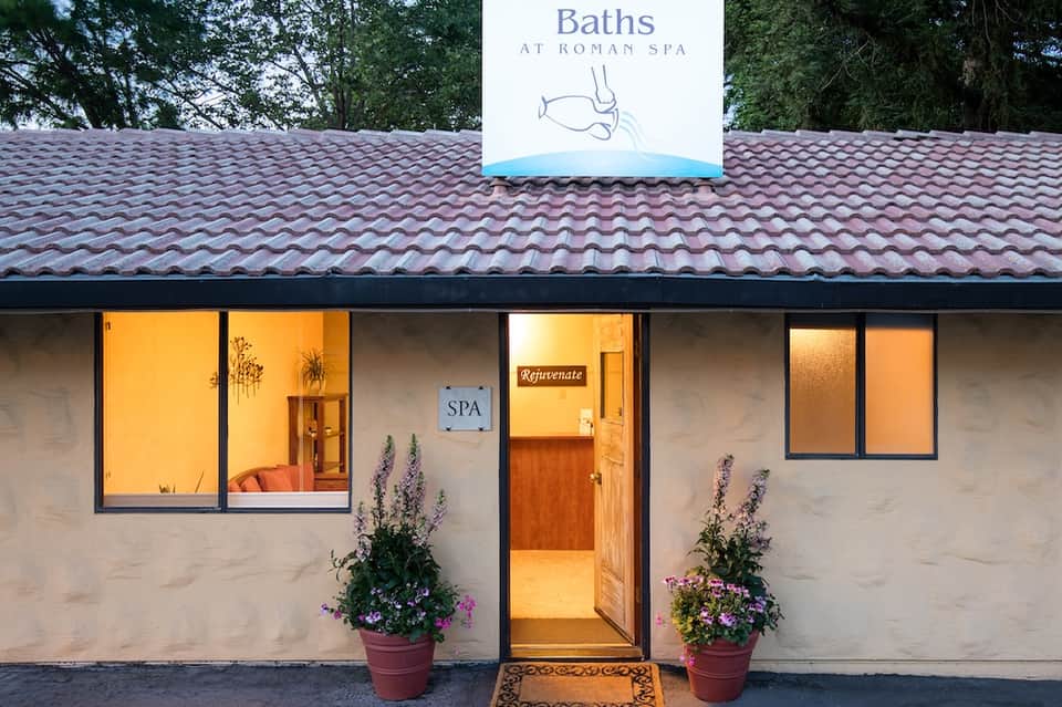 Roman Spa exterior with illuminated windows, purple flowers, and Baths signage on roof