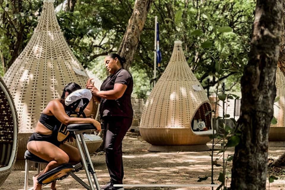 Outdoor fitness class with woman on stationary bike and instructor under woven pavilions in tree-shaded area
