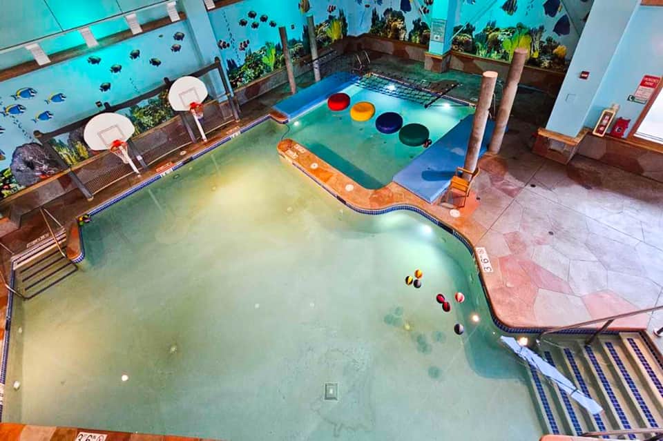 Indoor water park with turquoise pool, colorful floats, basketball hoop, and aquarium views