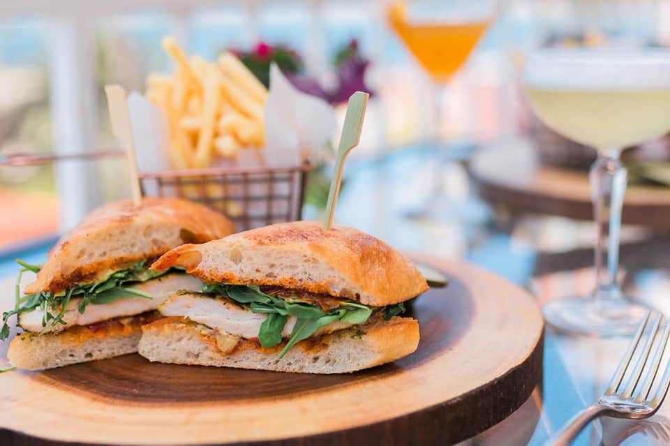 Gourmet sandwich with arugula and white cheese on wooden plate by poolside with cocktails and fries