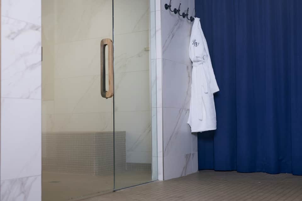 Modern spa shower area with marble tiles, glass enclosure, white robe hanging on wall, navy curtains