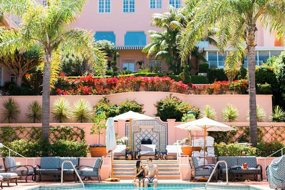 Elegant poolside courtyard with pink building, red flowers, palm trees, and lounge seating