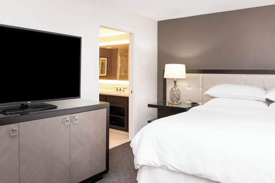 Hotel bedroom with TV, white bedding, gray headboard, and open bathroom door