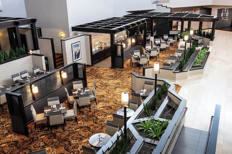 Modern event space with black pergolas, indoor seating areas, warm lighting, decorative plants, and patterned flooring