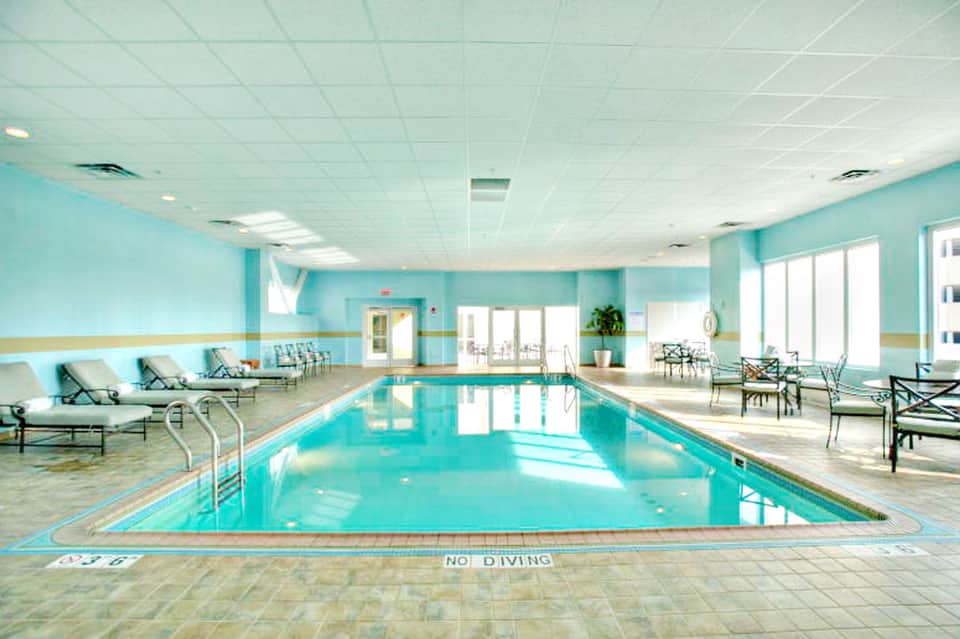 Indoor lap pool with turquoise water, lounge chairs, and large windows in a bright aquatic facility