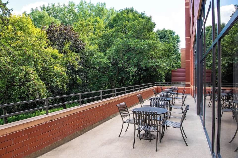 Outdoor terrace with dining tables, chairs, metal railings, and views of green trees