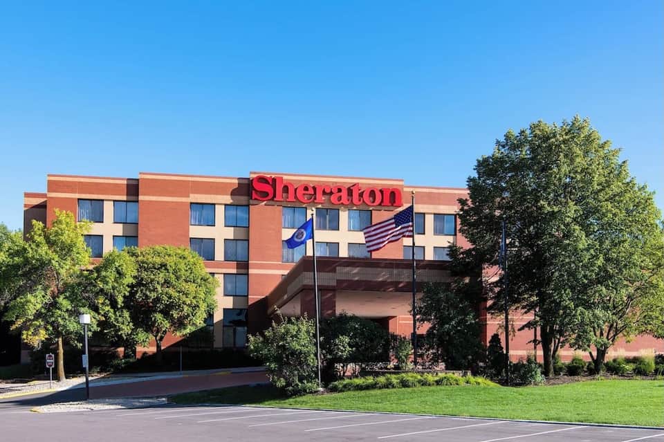 Red brick Sheraton hotel exterior with flags, manicured lawn, and surrounding trees