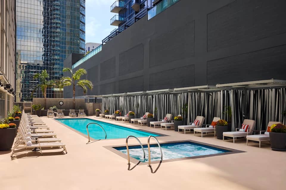 Urban hotel pool deck with lounge chairs, hot tub, and city building views with striped privacy screens