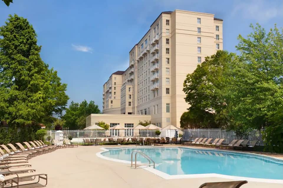 Multi-story hotel with outdoor swimming pool, lounge chairs, and mature trees on sunny day