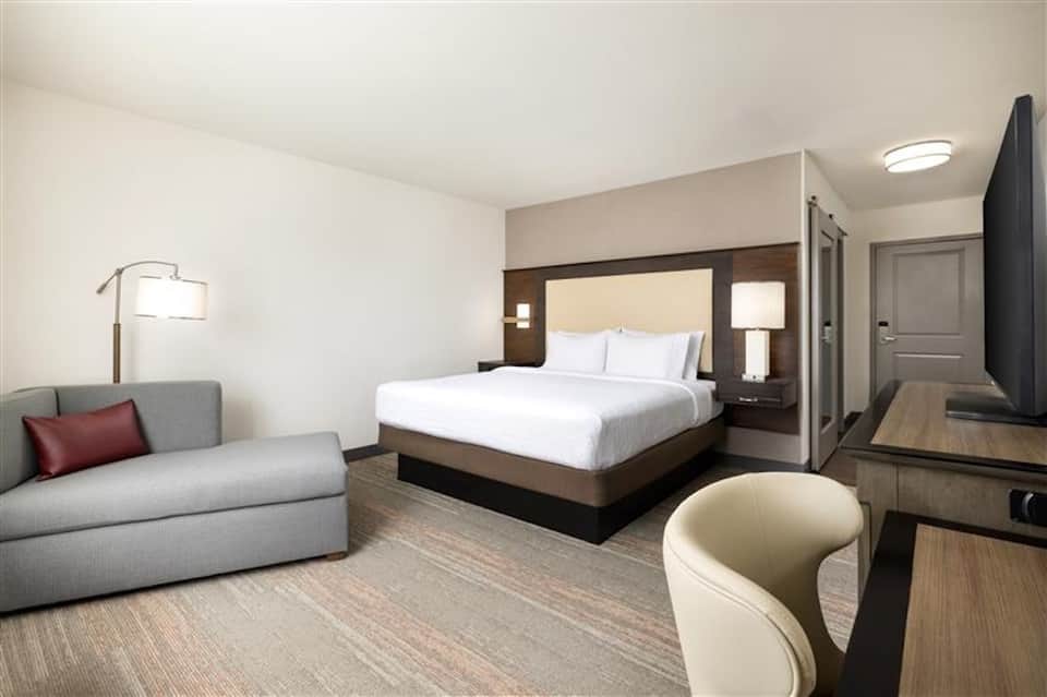 Modern hotel room with queen bed, gray sofa, wood headboard, and contemporary furnishings