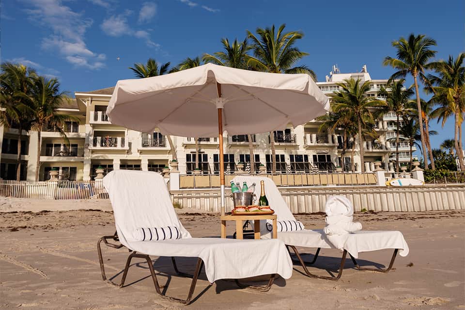Beach setup with white lounge chairs, large umbrella, champagne, and beachfront resort building