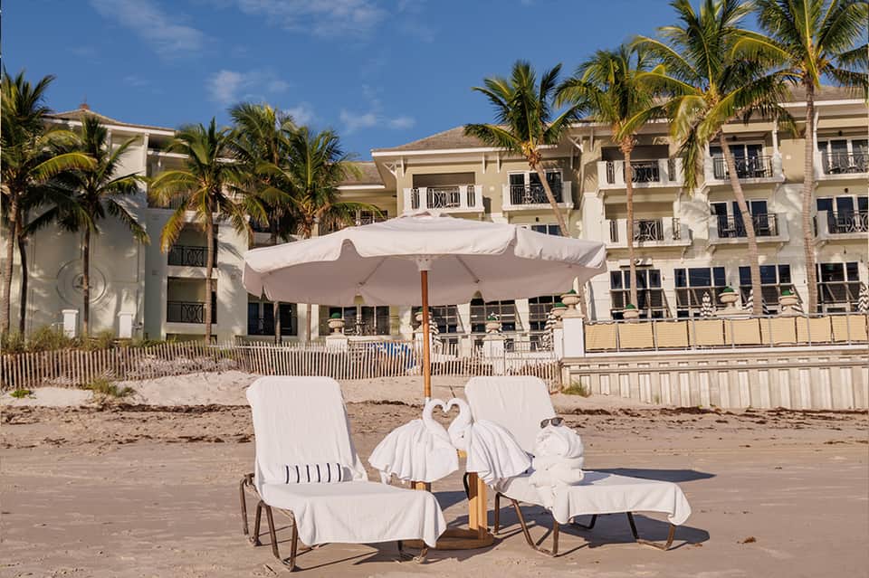 Beach loungers under white umbrella with resort buildings and palm trees, sandy beach setting
