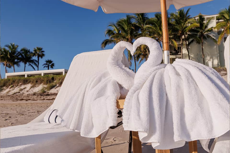Two white towels folded as swans on beach lounger with umbrella, palm trees and resort building behind