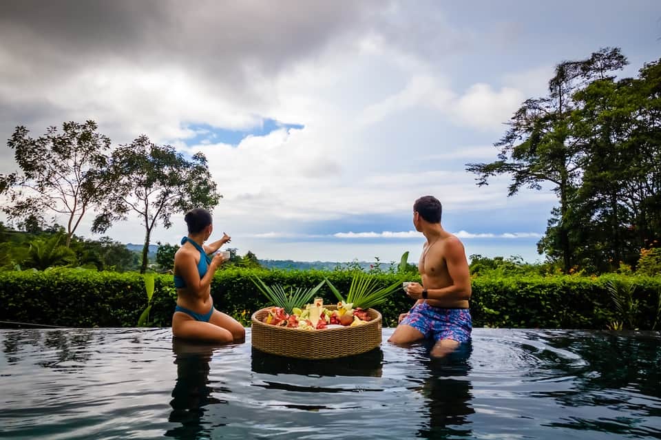 Two people in infinity pool sharing floating fruit basket with lush tropical landscape background