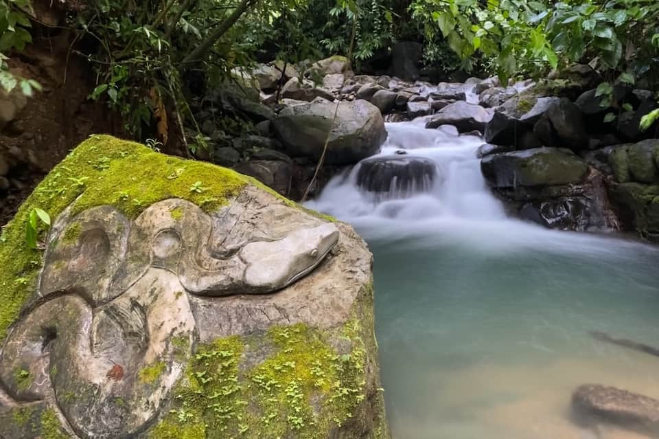 Moss-covered stone carving near cascading waterfall and emerald pool surrounded by lush tropical jungle vegetation