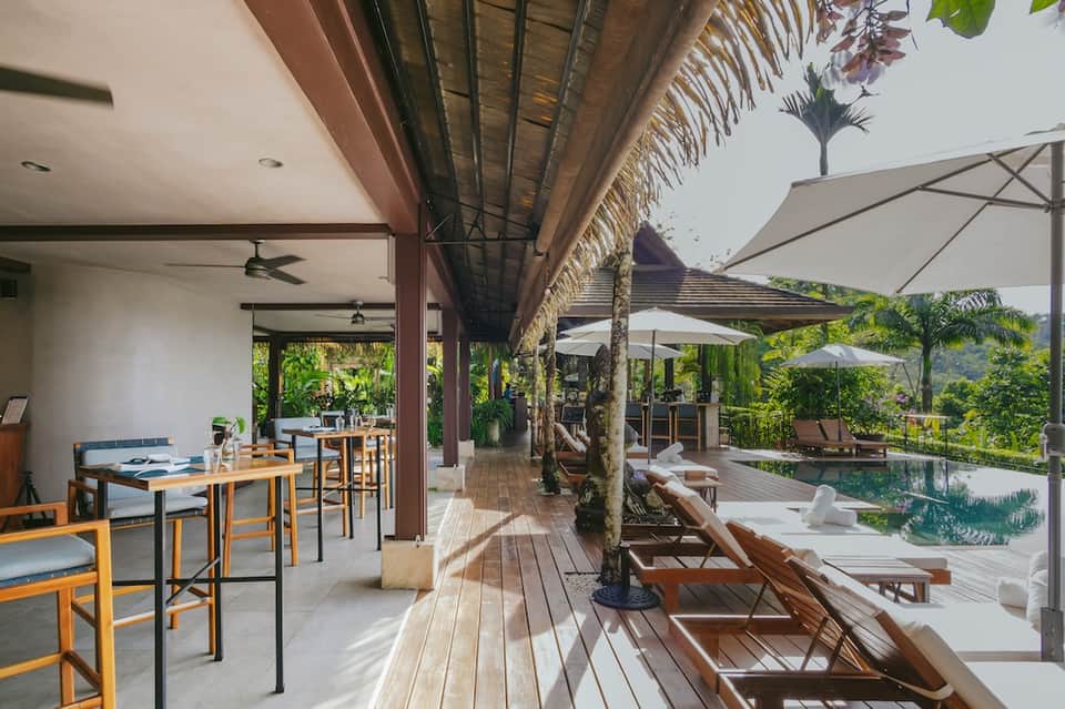 Modern resort terrace with wooden deck, dining area, lounge chairs, infinity pool, and jungle views