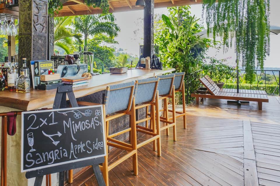 Outdoor hillside cafe with wooden bar counter, hanging plants, ocean views, and casual seating area