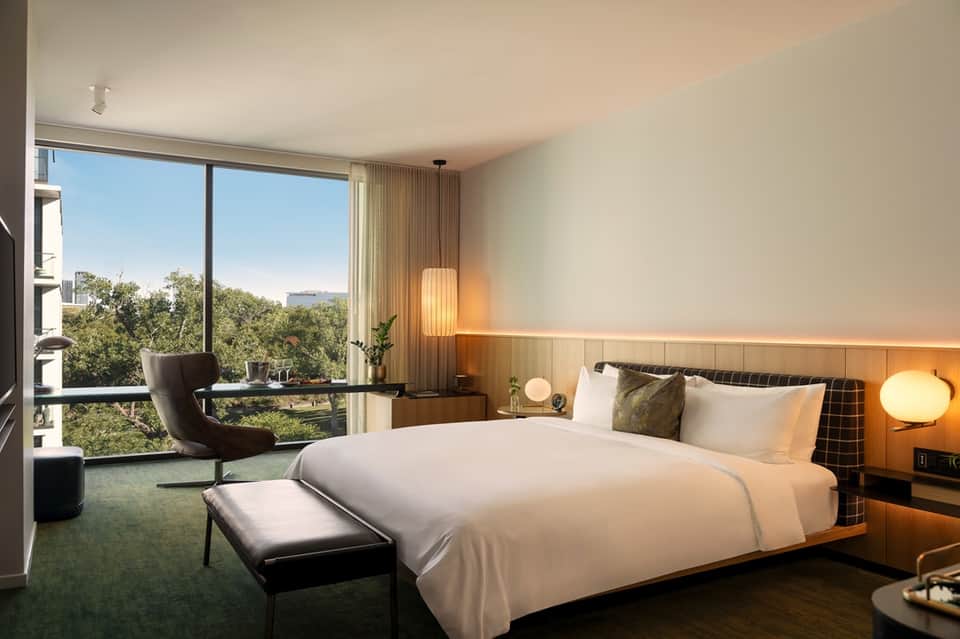 Luxury hotel bedroom with modern furnishings, floor-to-ceiling windows with nature views, and warm ambient lighting