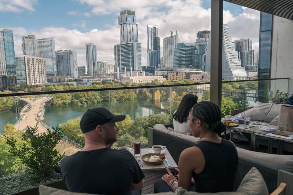 Three guests dining on high-rise restaurant balcony overlooking city skyline and river