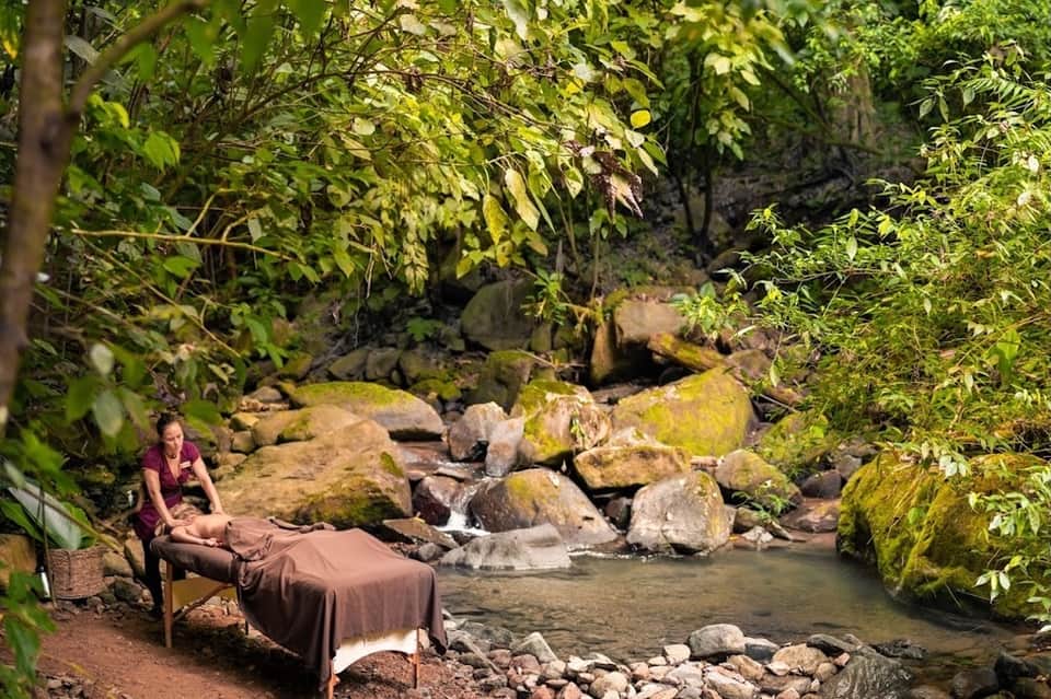 Woman receiving massage outdoors by stream with rocks and lush green forest surroundings