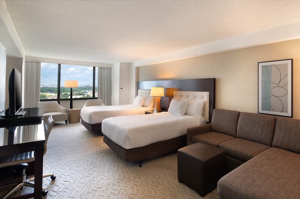 Modern hotel room with two beds, city view windows, and contemporary brown and beige decor