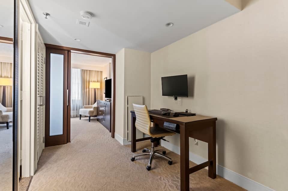 Hotel suite foyer with dark wood doors, desk workspace, and view into bedroom area with neutral color scheme.