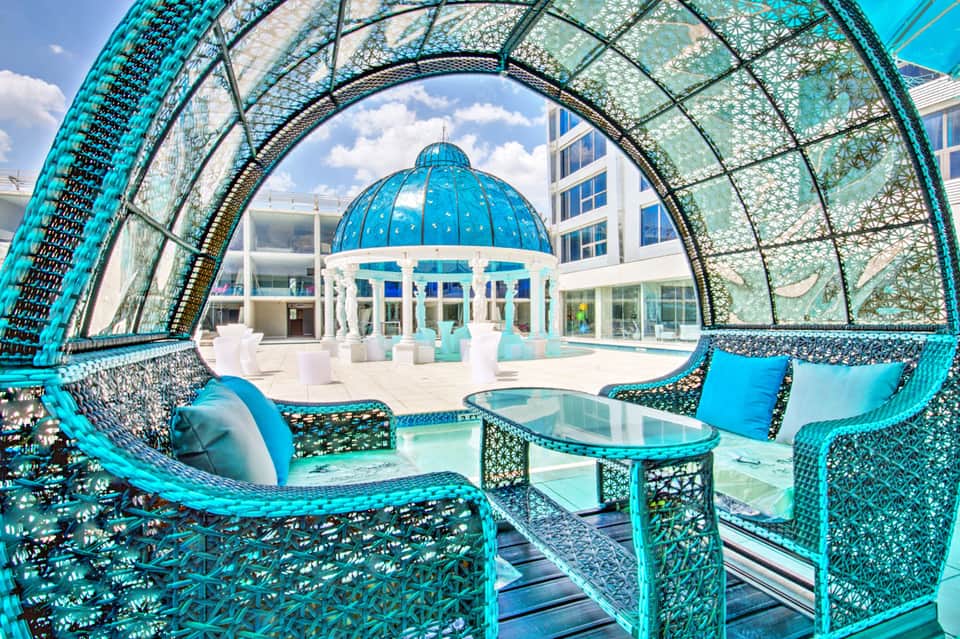 Turquoise woven lounge chairs under modern dome structure overlooking courtyard with white pavilion