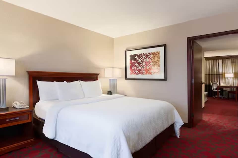 Hotel bedroom with queen bed, wood headboard, bedside lamps, and connecting suite