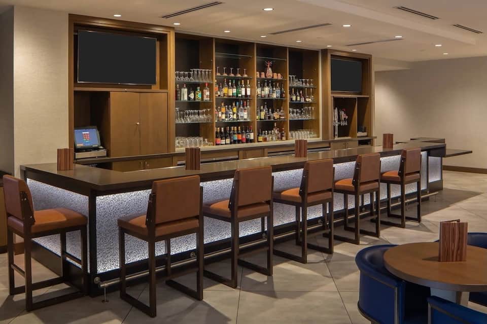 Hotel lounge with long wooden bar, leather chairs, backlit liquor bottles, and flat-screen TV