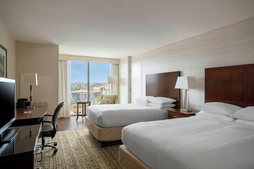 Modern hotel room with two queen beds, dark wood headboards, city view windows, and work desk with chair