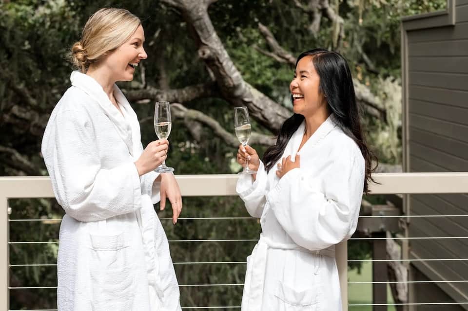 Two women in white spa robes toasting with champagne flutes in natural outdoor setting with trees