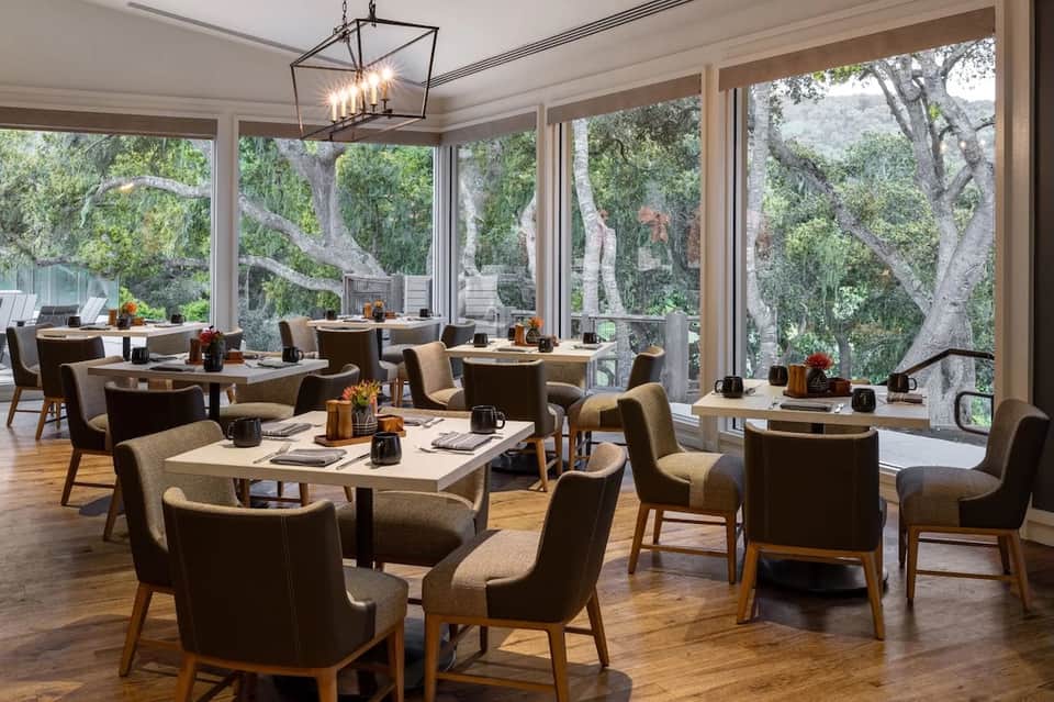 Upscale dining room with large windows overlooking trees, tan chairs, wooden tables, and pendant lighting