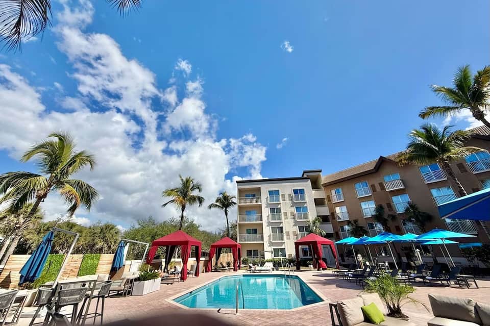 Resort pool area with colorful umbrellas, lounge chairs, palm trees, and hotel building