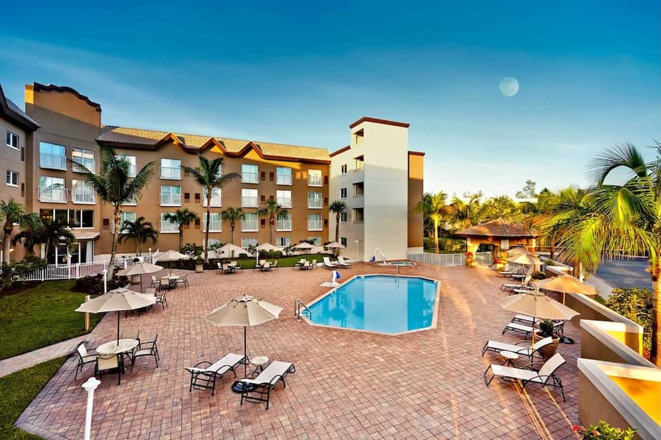 Resort exterior with multi-story building, pool area with loungers, umbrellas, and tropical landscaping