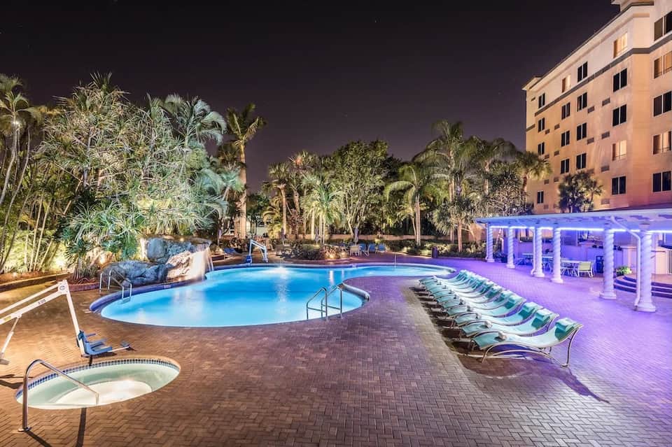 Illuminated resort pool at night with tropical landscaping, palm trees, and purple-lit cabana pavilion