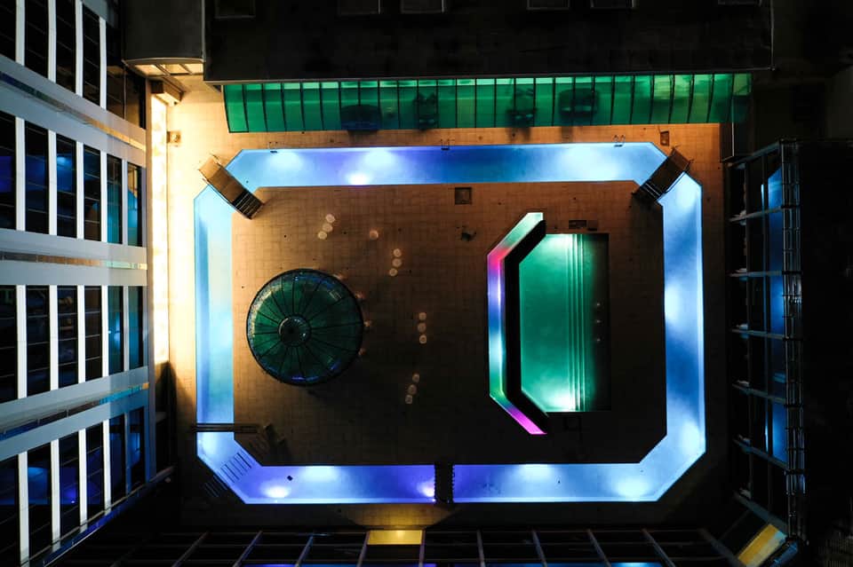 Overhead view of modern hotel courtyard with neon-lit green door, circular fan, and blue-lit floor borders at night