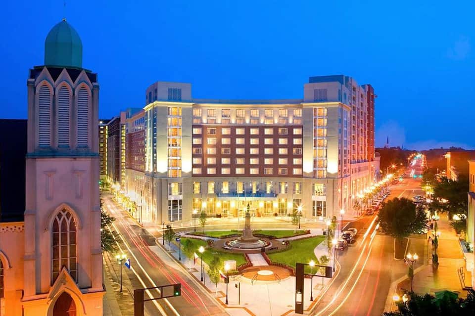 Illuminated modern hotel building at dusk with historic church tower and downtown cityscape