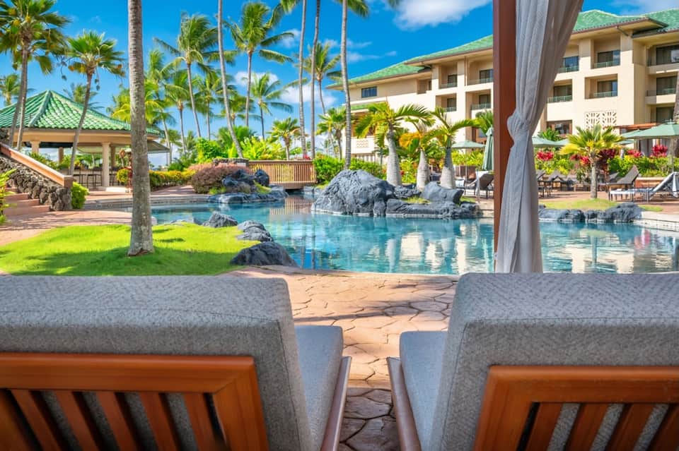 Tropical resort pool with palm trees, rock features, lounging chairs, and resort buildings