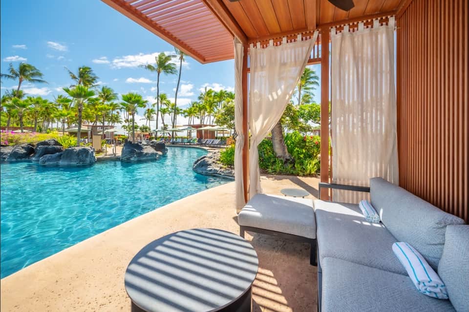 Tropical pool cabana with lounge seating, sheer curtains, and palm-lined lagoon view
