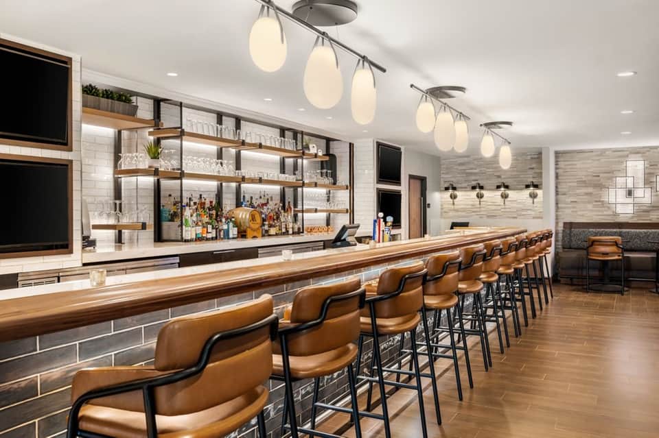 Modern hotel bar with wooden counter, leather bar stools, white tile backsplash, and pendant lighting